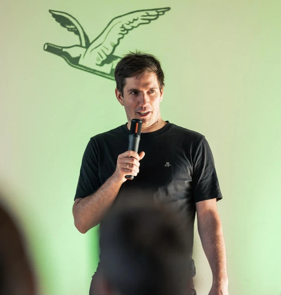Arturs Kruze speaking into a microphone in front of a pale green wall with a goose mural.