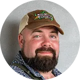 Bearded man wearing a beige cap with text and a blue checkered shirt, smiling at the camera.