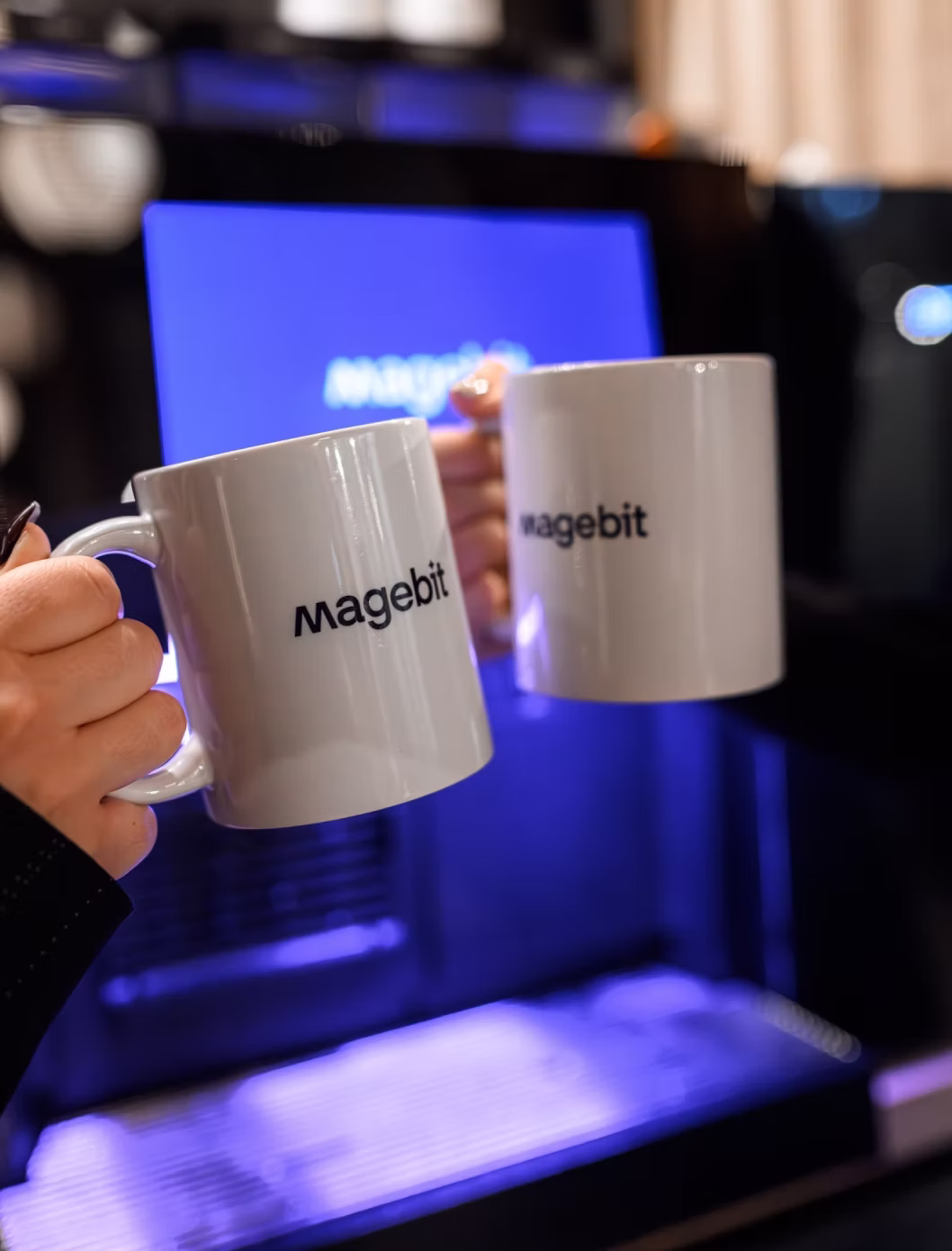 Two hands clinking white mugs with the word 'Magebit' in front of a blurred blue screen.