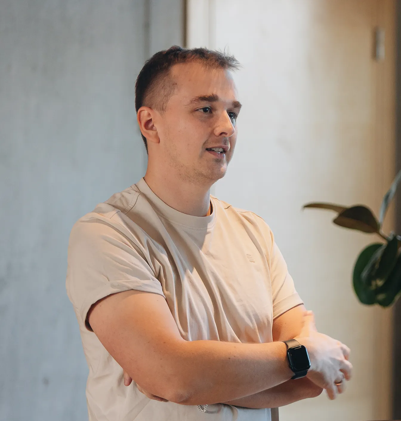 Man in beige t-shirt with arms crossed wearing a black smartwatch indoors near a plant.