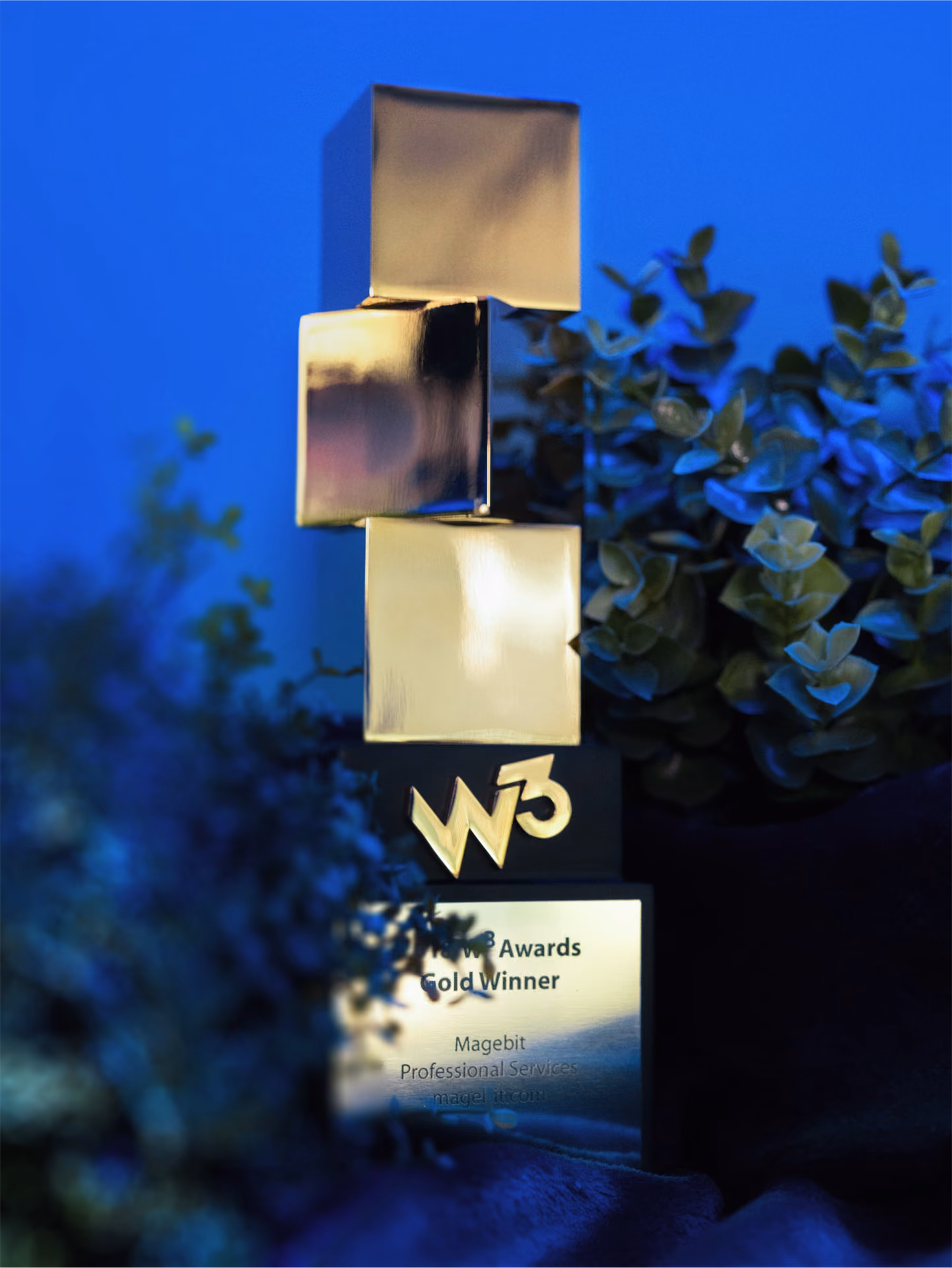 Gold W3 Awards trophy with three stacked cubes surrounded by green plants.