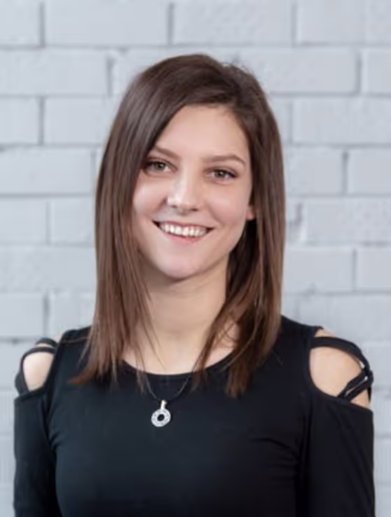 Smiling young woman with straight brown hair wearing a black cold-shoulder top and a silver necklace, standing against a gray brick wall.