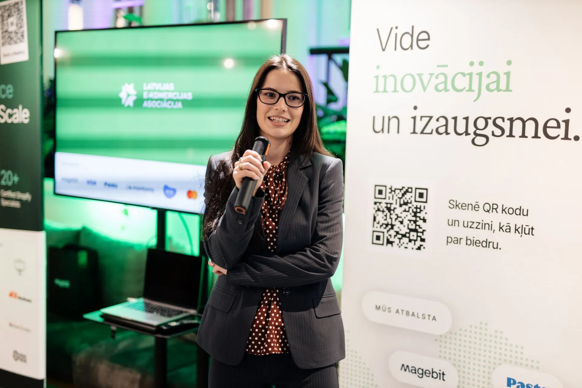 A LATEKA host welcoming guests with a microphone in front of the Latvian E-commerce Association banner