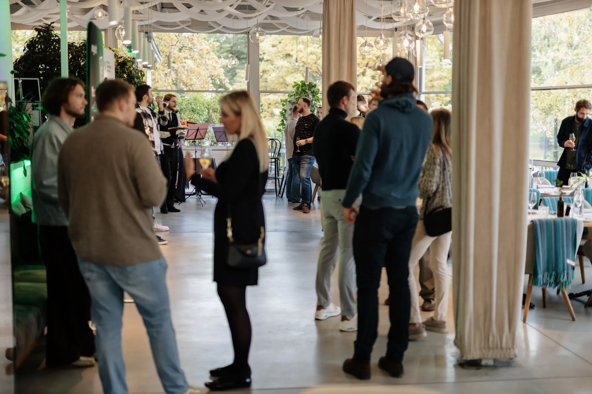 Guests gathering with drinks while a string duo plays by the windows at Kolonāde
