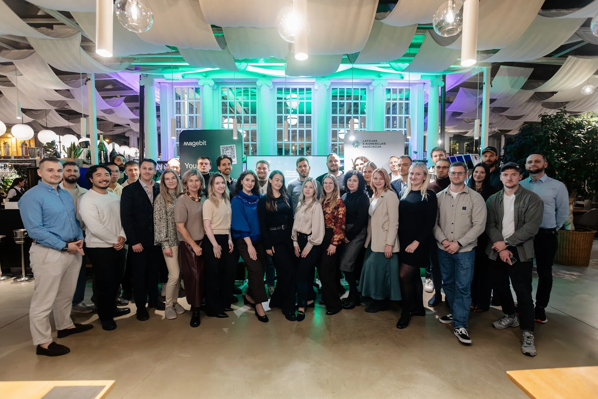 Group photo of guests and hosts at Kolonāde in front of the Magebit and LATEKA banners