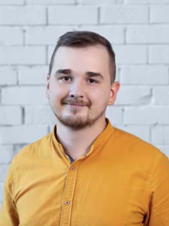 Young man with short brown hair and beard wearing a mustard yellow button-up shirt standing in front of a white brick wall.