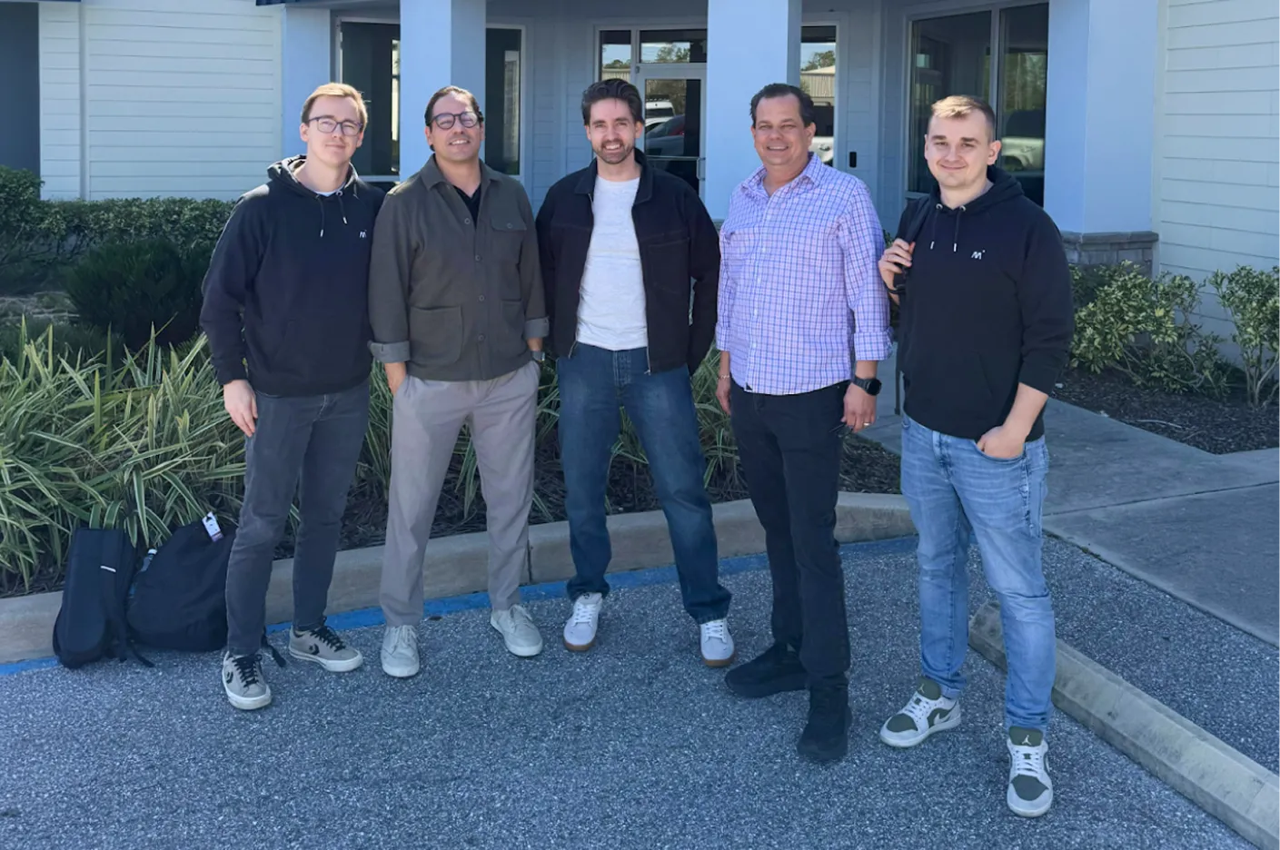 Five men standing outside a building on pavement with shrubs behind them, smiling at the camera.