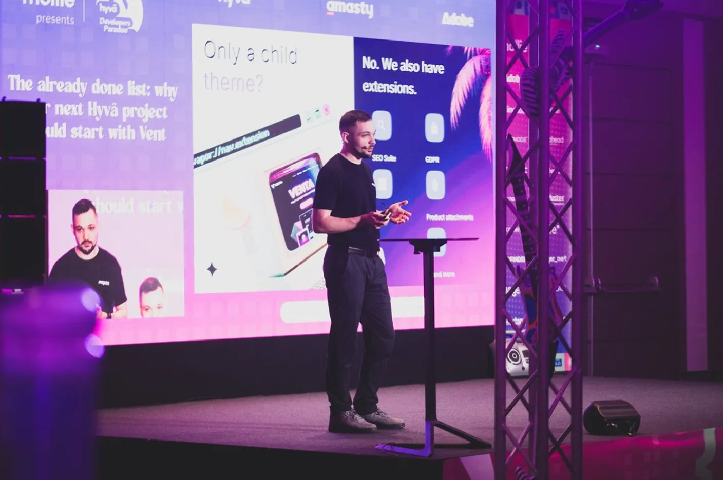 Man in black shirt giving a presentation on stage with a large screen behind him showing slides.