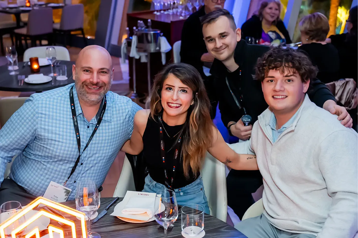 Four smiling people sitting and standing around a table in a restaurant setting.