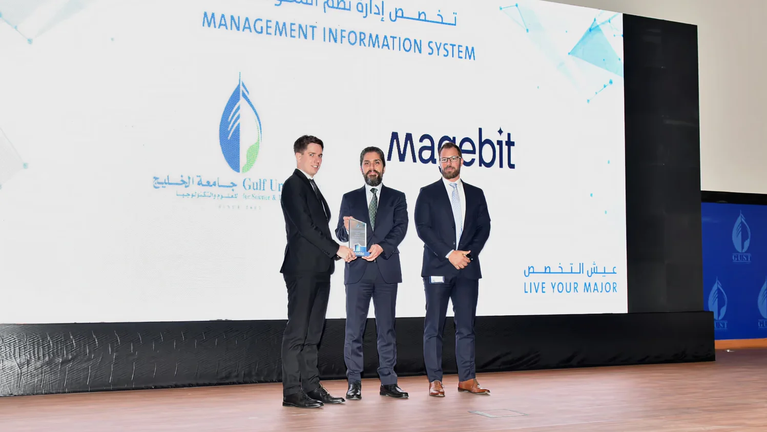 Magebit representatives receiving an award on stage at GUST's Live Your Major program in Kuwait, in front of the Magebit and Gulf University for Science and Technology logos