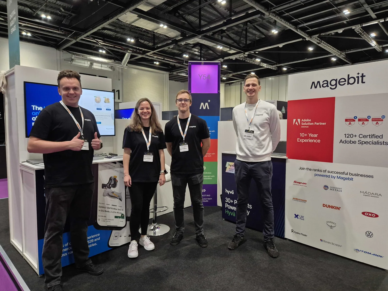Four Magebit team members at the Magebit stand at Digital Commerce Expo 2024, client logos and Adobe Solution Partner panel behind them