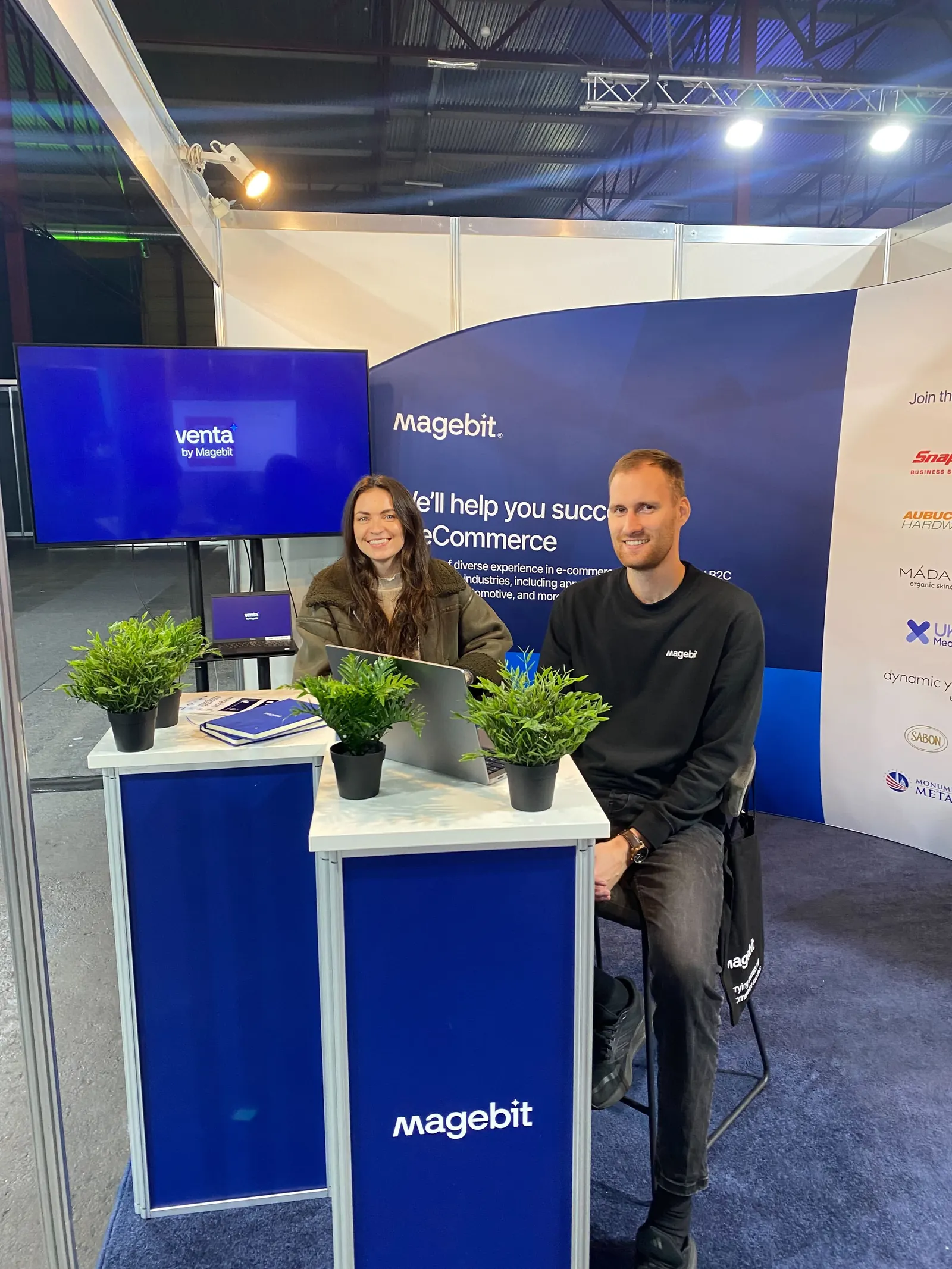 Two Magebit team members seated at the Magebit booth at RIGA COMM 2025, with Venta by Magebit on the screen behind them