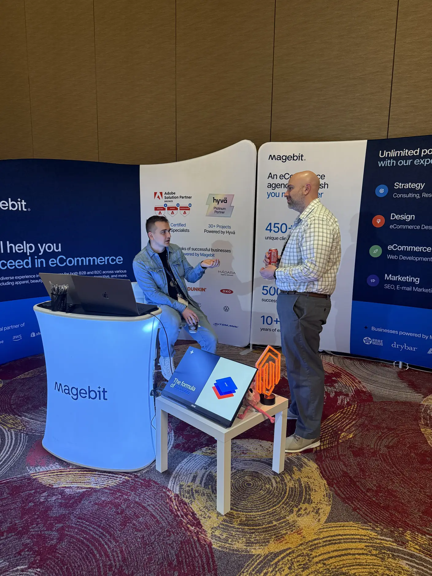 The Magebit booth at Meet Magento Florida 2025, with a laptop demo stand and two people talking