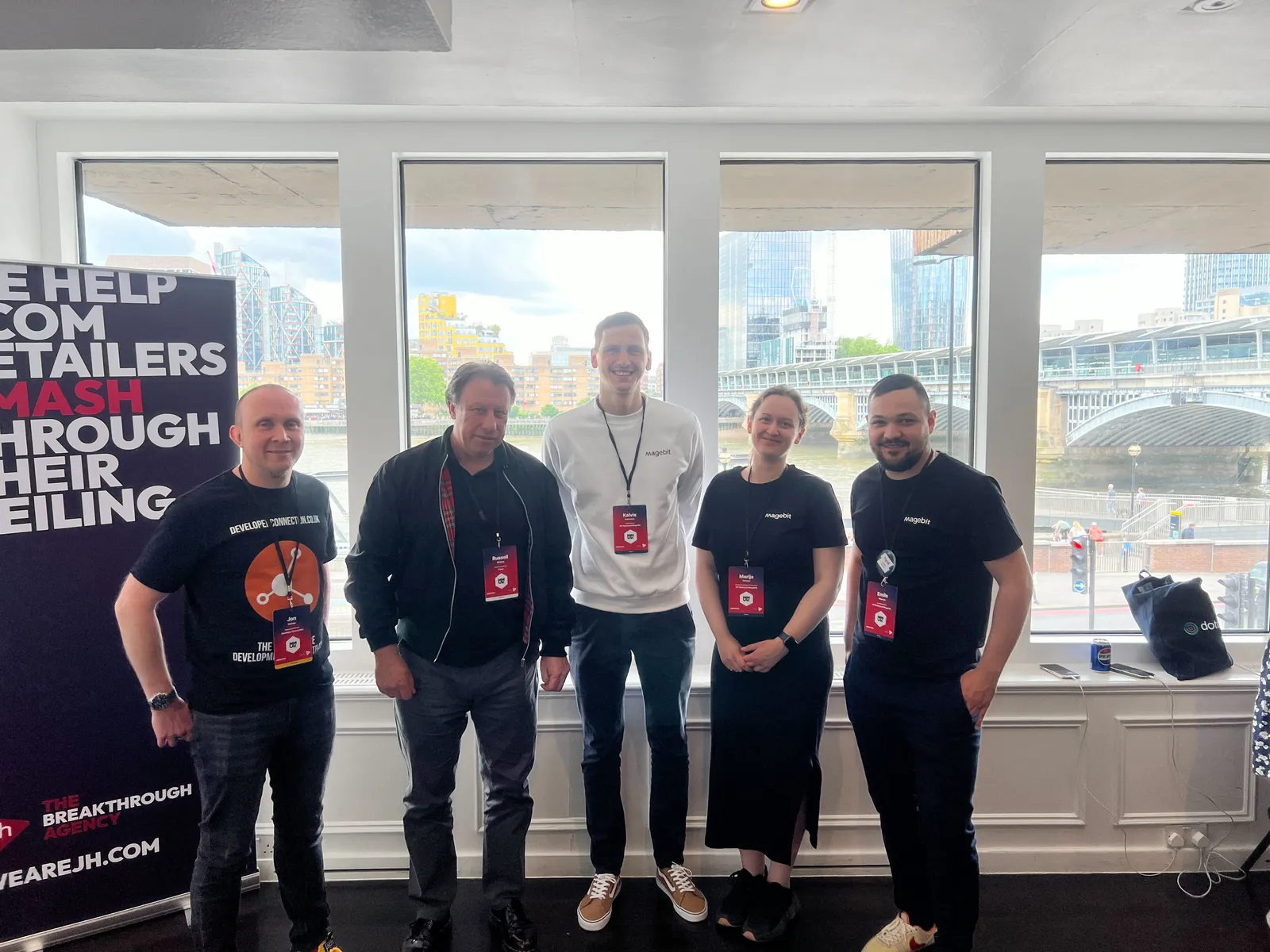 Magebit team with conference badges beside a JH banner at The Mermaid, Meet Magento UK 2024