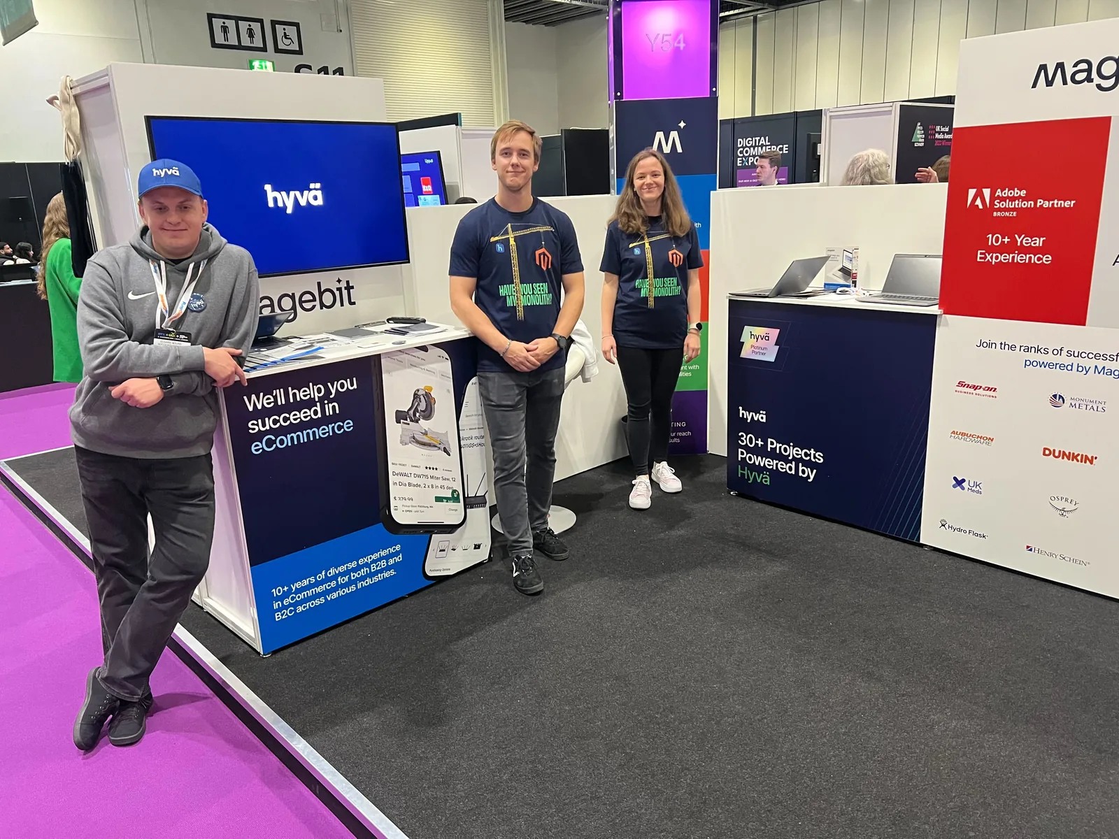 Three Magebit team members at the Magebit stand at Digital Commerce Expo 2024, with Hyvä Platinum Partner and Adobe Solution Partner panels