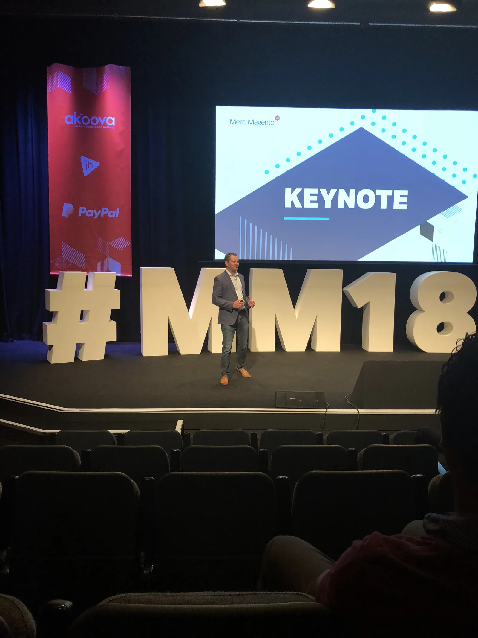 Speaker in front of the giant #MM18 letters during a keynote at Meet Magento UK 2018, with Akoova, JH and PayPal banners