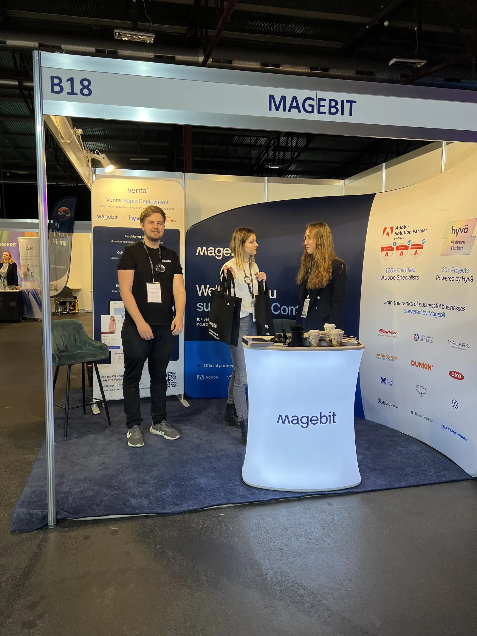 Three Magebit team members at stand B18 at RIGA COMM 2024, with Venta, Adobe Solution Partner and Hyvä Platinum Partner panels behind them