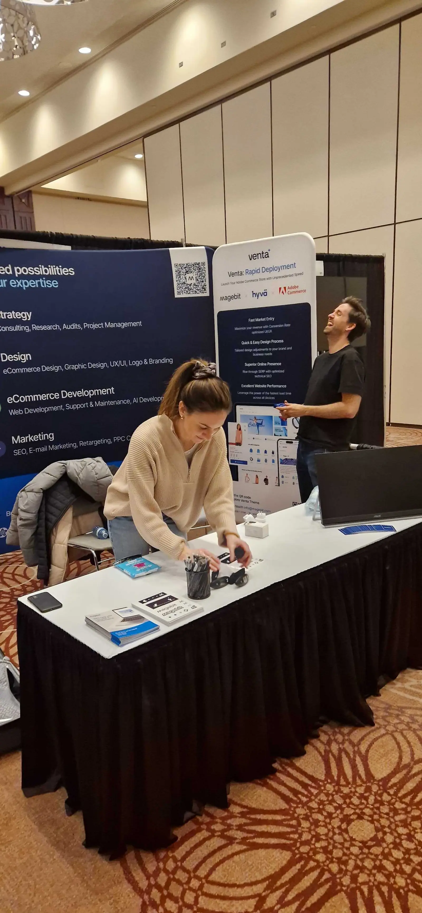 A Magebit team member arranging brochures on the booth table beside the Venta, Hyvä and Adobe Commerce roll-up at DX3 Canada 2025