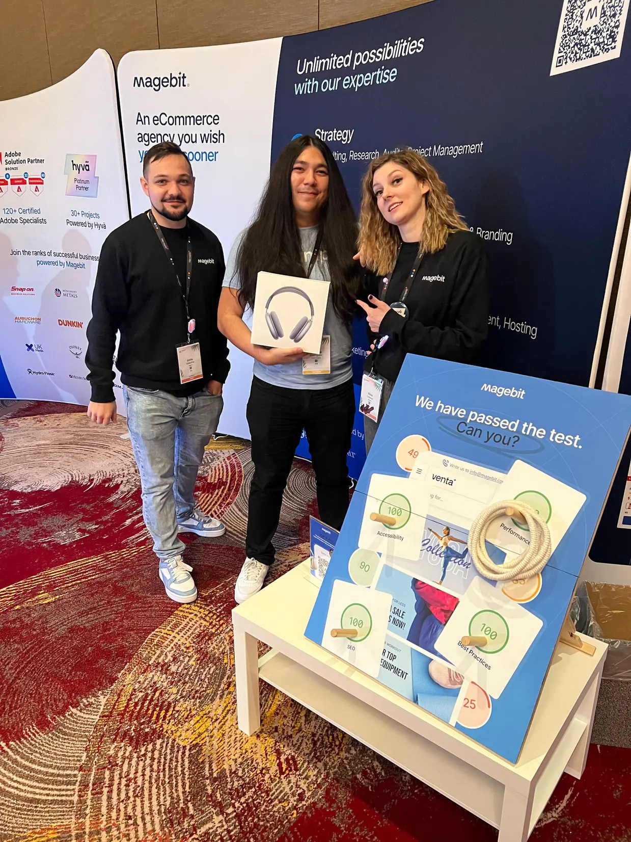 Three people at the Magebit booth at Meet Magento Florida 2025, one holding a pair of prize headphones