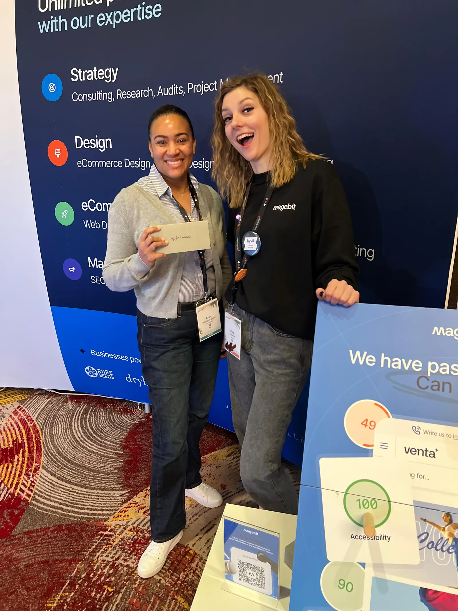 Two people posing at the Magebit booth at Meet Magento Florida 2025 beside the "We have passed the test" display