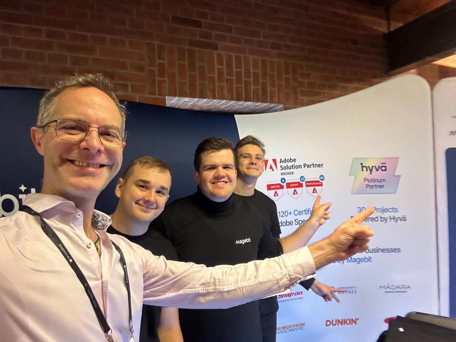 Magebit team taking a selfie with a visitor in front of the Magebit booth wall at Meet Magento Poland 2024