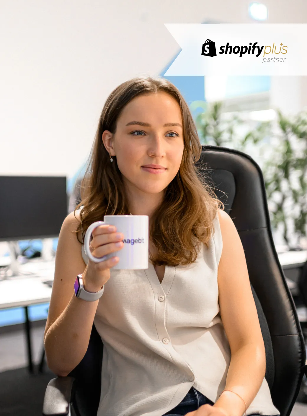 Woman sitting in an office chair holding a Magebit mug with Shopify Plus partner logo above.