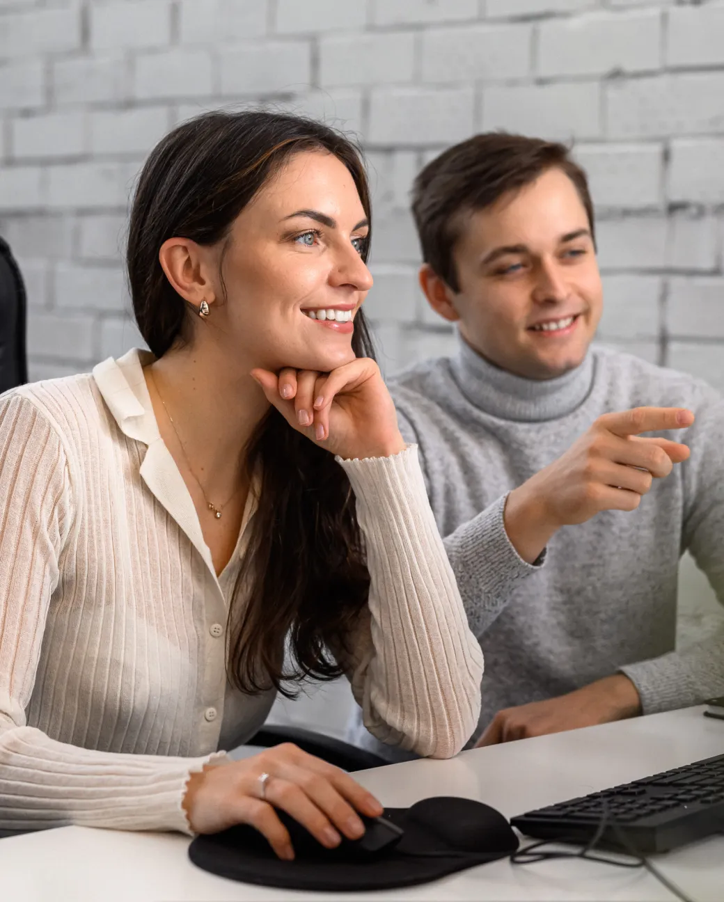 Smiling woman using computer mouse as man beside her points at the screen in a light office setting.